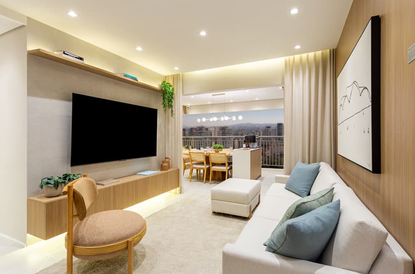 Sala de estar com painel para TV em tons de madeira, sofa claro e vista para a varanda.